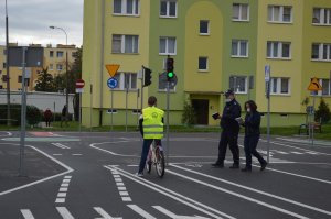 Nastolatek jedzie rowerem po miasteczku rowerowym, policjant i osoba cywilna przyglądają się.