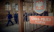 Zdjęcie z wejścia na teren szkoły policji. Z prawej strony tablica informacyjna &quot;Szkoła Policji w Słupsku&quot;