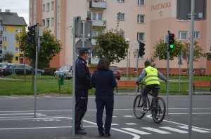 Nastolatek jedzie rowerem po miasteczku rowerowym, policjant i osoba cywilna przyglądają się.
