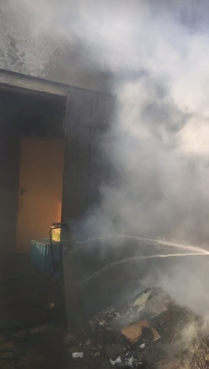 Gaszenie palących się drzwi od pomieszczenia gospodarczego za pomocą węża ogrodowego. Widać unoszącą się łunę dymu.