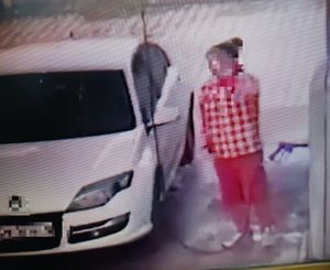 zdjęcie kolorowe, niewyraźne, sprawca kradzieży lancy ubrany w czerwono białe barwy i obok biały samochód osobowy