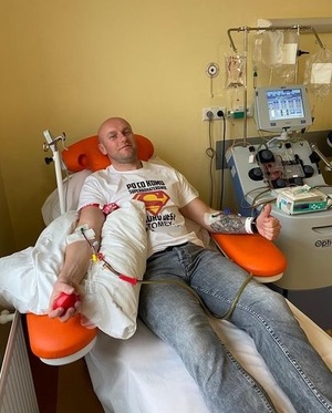Zdjęcie kolorowe zrobione w szpitalu, w pokoju zabiegowym. Widoczny biały mężczyzna w średnim wieku na łóżku podłączony do aparatury pobierającej szpik kostny.