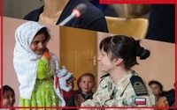 Zdjęcie kolorowe, z lewej strony kilkuletnia dziewczynka w zielonej sukience i chuście na głowie, z prawej strony kobieta w wojskowym mundurze spogląda na dziewczynkę