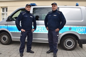 Zdjęcie kolorowe. Dwóch policjantów w mundurach przed radiowozem