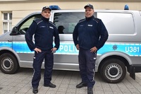 Zdjęcie kolorowe. Dwóch policjantów w mundurach przed radiowozem