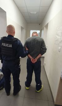Zdjęcie wykonane na korytarzu, dwóm osobom od tyłu. Policjant wykonuje czynności z podejrzanym.