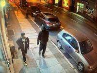 Zapis kolorowy z nocy wykonany przez kamerę monitoringu. Widok ulicy, po chodniku porusza się dwóch zamaskowanych mężczyzn ubranych na czarno, przy chodniku stoją zaparkowane auta.