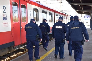 Trzech funkcjonariuszy SOP i dwóch policjantów idą wzdłuż czerwonego pociągu, który zatrzymał się przy peronie.