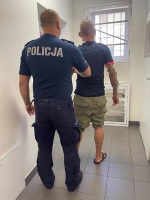 Policjant wykonuje czynności z zatrzymanym mężczyznę. Funkcjonariusz i zatrzymany od tyłu.