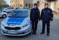 Policjant i policjantka w granatowych mundurach, przy radiowozie.