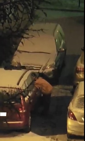 Na zdjęciu widoczna sylwetka mężczyzny, który zagląda przez wybitą szybę do auta. Obok w pokrywie śnieżnej zaparkowane są inne pojazdy.