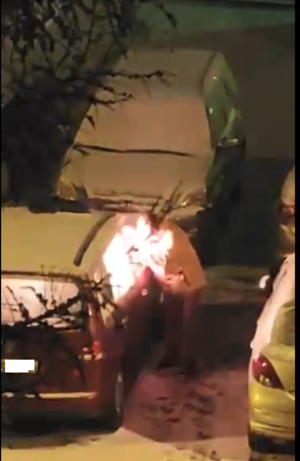 Na zdjęciu widoczna sylwetka mężczyzny, który zagląda przez wybitą szybę do auta. Widać pożar w środku samochodu. Obok w pokrywie śnieżnej zaparkowane są inne pojazdy.