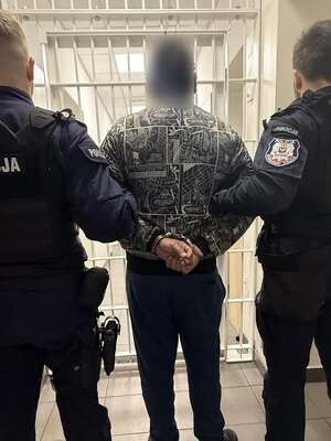 Policjanci z lewej i prawej strony, w środku mężczyzna z który jest w trakcie doprowadzania do radiowozu. Zdjęcie wykonane od tyłu.